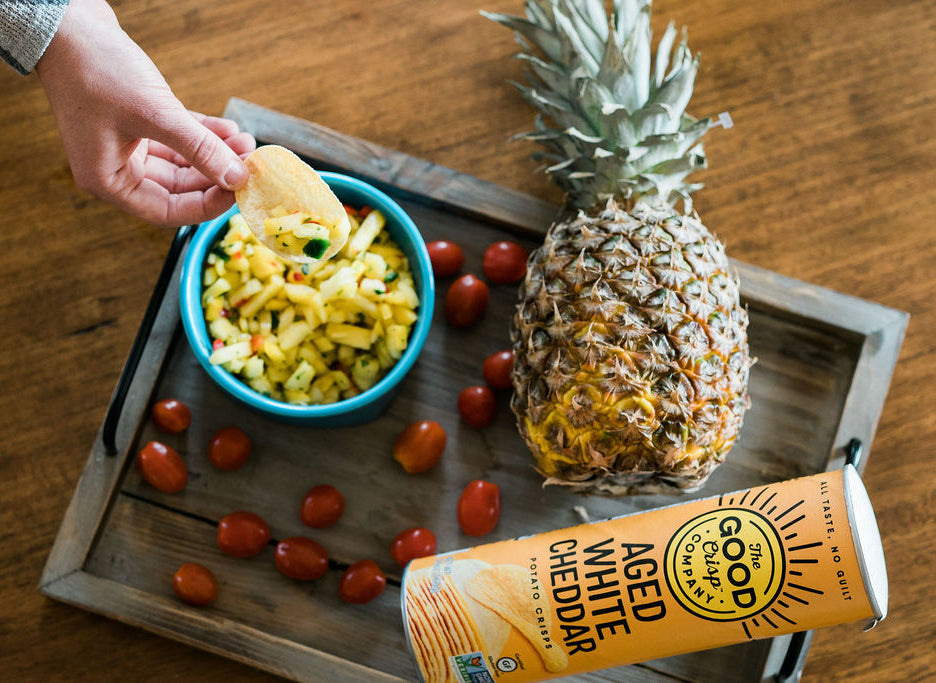 A bowl of yellow pineapple salsa, next to a pineapple, some tomatoes and a tube of Aged white cheddar chips. Someone is scooping some salsa onto a chip